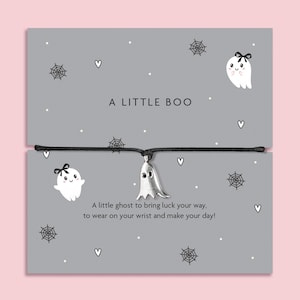 Op de afbeelding: Een zilverkleurige spookbedel aan een zwart koordarmbandje, gepresenteerd op een grijze kaart met illustraties van spinnenwebben en spookjes, met de tekst A LITTLE BOO en A little ghost to bring luck your way, to wear on your wrist and make your day!