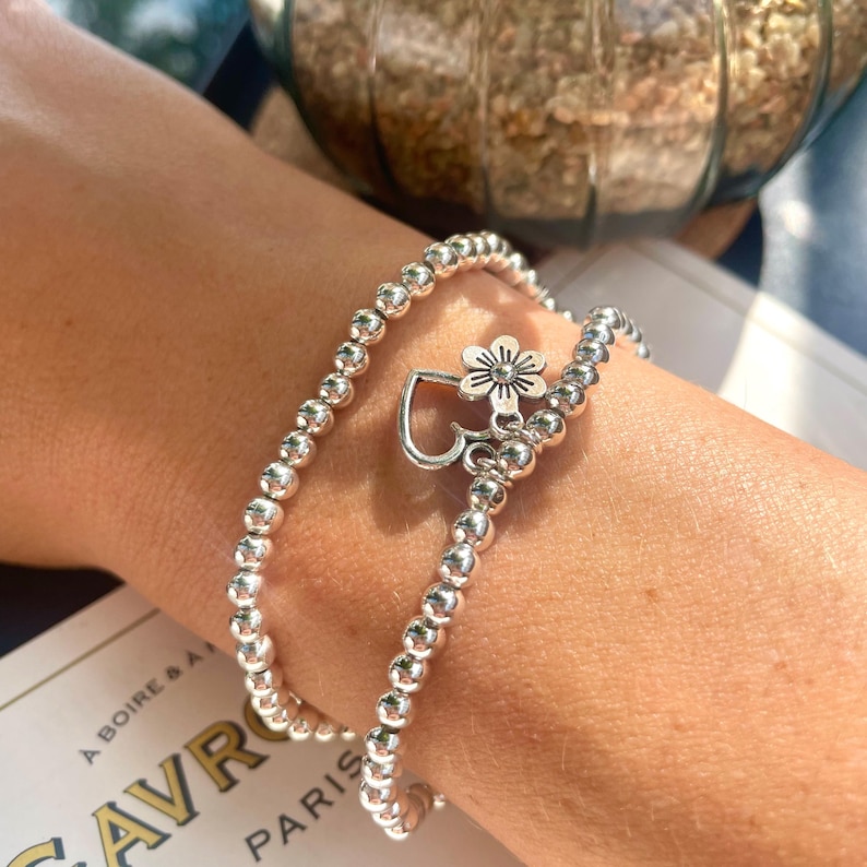 May include: Two silver beaded bracelets with a silver charm featuring a heart and a flower. The bracelets are stacked on a person's wrist.