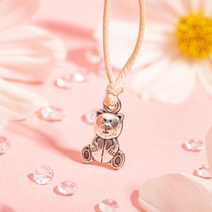 May include: A silver-colored teddy bear pendant necklace with a beige cord. The bear is sitting with its arms at its sides. The background is pink with white flowers and clear crystals.