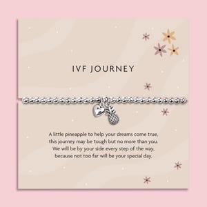 Może przedstawiać: Srebrna bransoletka z koralików z zawieszką w kształcie serca i ananasa, prezentowana na jasnoróżowej karcie. Na karcie widnieje napis "IVF JOURNEY" i zawiera wspierającą wiadomość. Kwiatowe akcenty zdobią kartę.