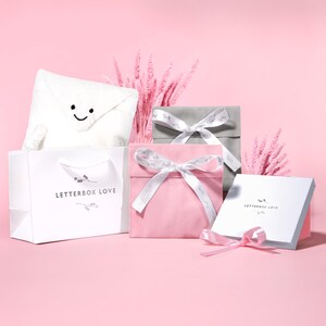 May include: A collection of gift boxes and a shopping bag on a pink background. The boxes are in pink, gray, and white, tied with white ribbons. A white bag with "LETTERBOX LOVE" printed on it holds a plush envelope with a smiling face.