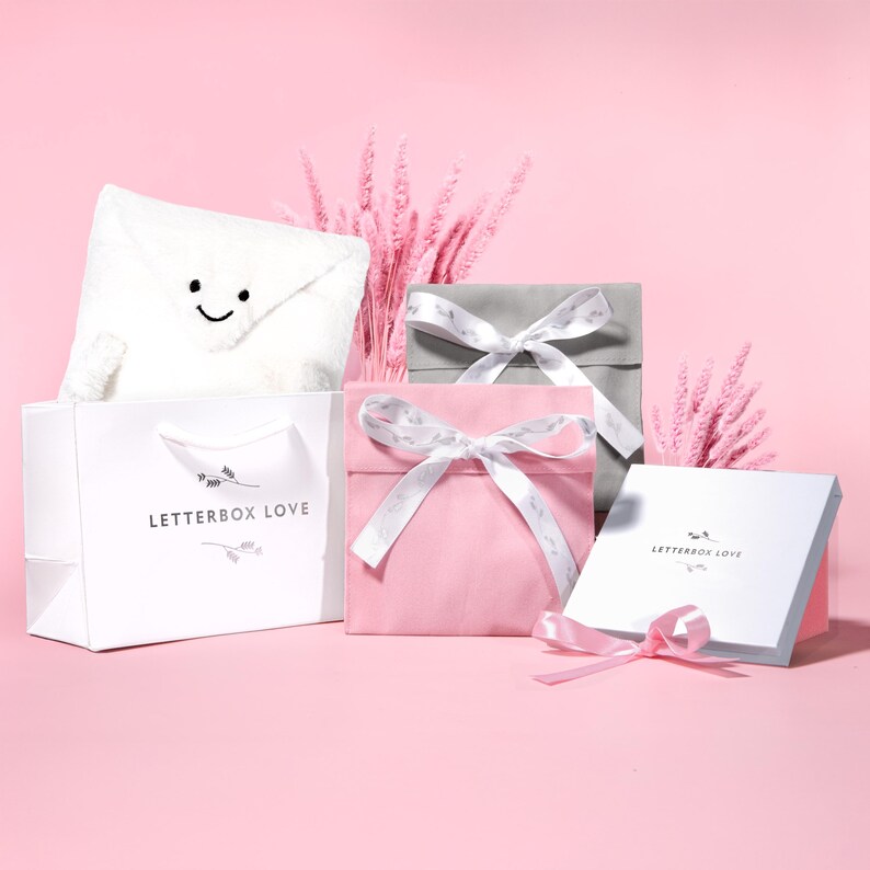 May include: A collection of gift boxes and a shopping bag on a pink background. The boxes are in pink, gray, and white, tied with white ribbons. A white bag with "LETTERBOX LOVE" printed on it holds a plush envelope with a smiling face.