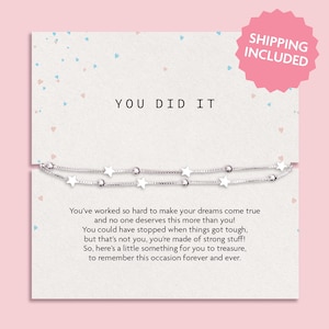 Op de afbeelding: Een zilveren armband met sterbedels wordt gepresenteerd op een witte kaart met de woorden "YOU DID IT". De kaart heeft een roze en blauw hartpatroon. De tekst hieronder luidt: "Je hebt zo hard gewerkt...om deze gelegenheid voor altijd te herinneren."