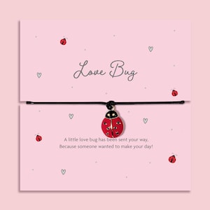 Puede incluir: Una tarjeta rosa con las palabras "Love Bug" en escritura cursiva. Una pulsera de hilo negro con un dije de mariquita roja. La tarjeta tiene pequeñas ilustraciones de mariquitas y corazones, con el texto "A little love bug has been sent your way, Because someone wanted to make your day!"
