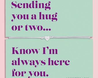 May include: A light green card featuring the purple text "Sending you a hug or two..." and "Know I'm always here for you." A thin white string bracelet with a small silver heart charm is positioned horizontally across the center of the card.