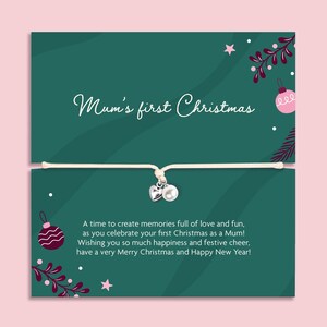 May include: A string bracelet with a silver heart charm and a white pearl bead, attached to a dark green card featuring the text Mum's first Christmas and a holiday message, decorated with pink ornaments, stars, and dark red foliage illustrations.