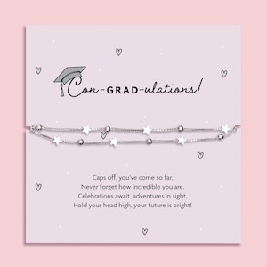 Pode incluir: Uma pulseira prateada com berloques em forma de estrela e pequenas contas é exibida em um cartão rosa. O cartão tem o texto "Con-GRAD-ulations!" e uma ilustração de um chapéu de formatura. Uma mensagem de parabéns e ilustrações de corações também estão presentes.