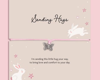 May include: A light pink string bracelet featuring a silver-toned butterfly charm, presented on a beige card with white rabbit illustrations, pink flowers, and the text "Sending Hugs" and "I'm sending this little hug your way, to bring love and comfort to your day."