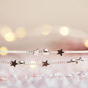 May include: A silver-toned box chain bracelet featuring alternating small star-shaped charms and smooth round spherical beads. The delicate metallic chain is arranged in two parallel lines, showcasing a polished finish and consistent spacing.