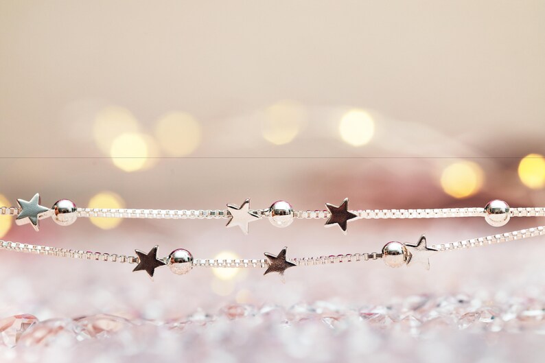 May include: A silver-toned box chain bracelet featuring alternating small star-shaped charms and smooth round spherical beads. The delicate metallic chain is arranged in two parallel lines, showcasing a polished finish and consistent spacing.