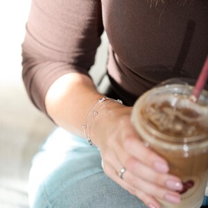 May include: A delicate silver-toned chain bracelet featuring small, evenly spaced metallic beads and clear, sparkling stones. The bracelet is designed with a thin, layered appearance and a secure clasp, resting on a wrist alongside a simple silver ring.