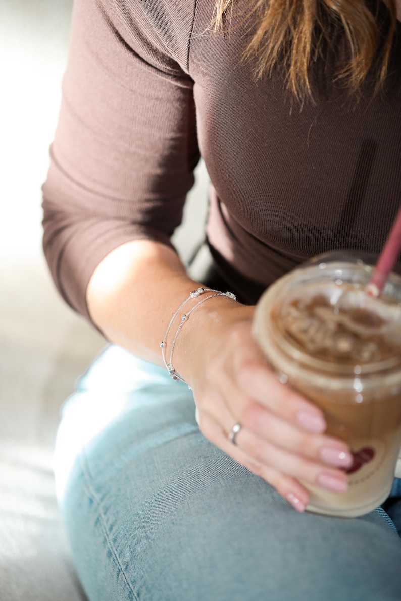 May include: A delicate silver-toned chain bracelet featuring small, evenly spaced metallic beads and clear, sparkling stones. The bracelet is designed with a thin, layered appearance and a secure clasp, resting on a wrist alongside a simple silver ring.