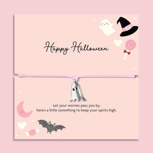 Op de afbeelding: Een zilveren spookbedel aan een lichtroze koord, gepresenteerd op een lichtroze kaart met de tekst Happy Halloween en Let your worries pass you by, here's a little something to keep your spirits high, met illustraties van een spook, heksenhoed, lolly, maan, hart en vleermuis.