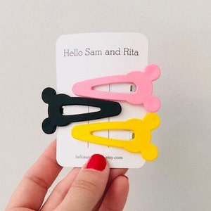 May include: Three plastic hair clips in the shape of a bear's head. The clips are pink, yellow, and black. The clips are on a white card with the text "Hello Sam and Rita" printed on it.