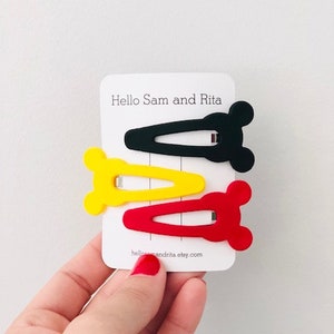 May include: Three plastic hair clips in the shape of a cartoon mouse head. The clips are red, yellow, and black.