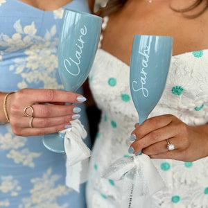 Puede incluir: Dos copas de champán azul claro con cintas blancas y nombres personalizados "Claire" y "Sarah". Las copas están sostenidas por dos personas y la cinta de la copa derecha tiene el texto "Sarah's Bridal Shower" impreso.