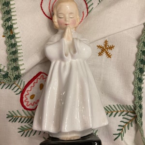 Puede incluir: Una figurita de porcelana blanca de un niño rezando con las manos juntas. La figurita lleva una túnica blanca y tiene el pelo rubio. La figurita está de pie sobre una base negra.
