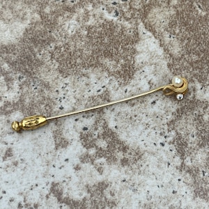 May include: A gold-toned stick pin with a pearl and a small clear crystal accent. The pin has a decorative end and a simple, elegant design. The background is a neutral, textured surface.