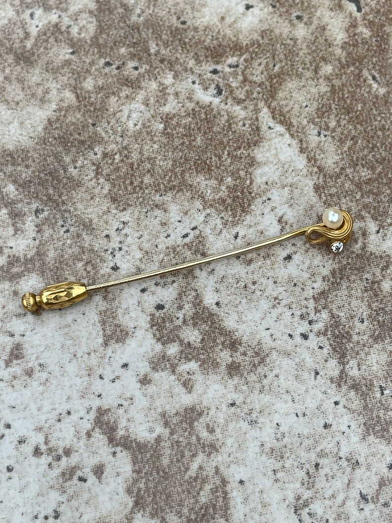 May include: A gold-toned stick pin with a pearl and a small clear crystal accent. The pin has a decorative end and a simple, elegant design. The background is a neutral, textured surface.
