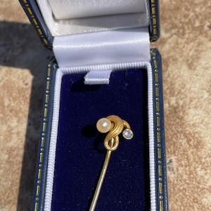 May include: A gold-toned stick pin with a pearl and a small, clear gemstone, presented in a blue jewelry box with a white satin lining. The pin's design features a swirling top.