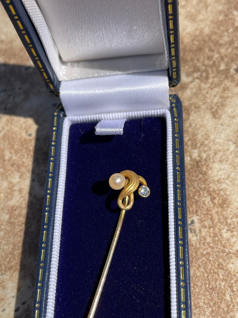 May include: A gold-toned stick pin with a pearl and a small, clear gemstone, presented in a blue jewelry box with a white satin lining. The pin's design features a swirling top.