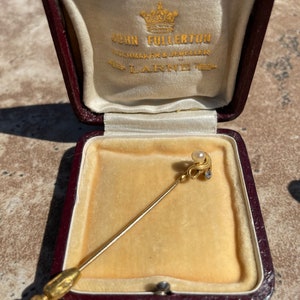 May include: A gold-colored stick pin with a pearl and small diamond accents, displayed in a burgundy jewelry box with a cream-colored satin lining. The box is open, revealing the pin against a yellow velvet interior.