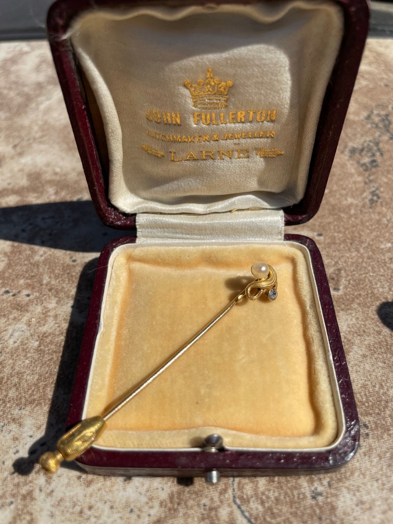 May include: A gold-colored stick pin with a pearl and small diamond accents, displayed in a burgundy jewelry box with a cream-colored satin lining. The box is open, revealing the pin against a yellow velvet interior.