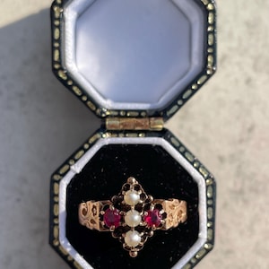 Puede incluir: Anillo dorado con tres perlas y dos piedras rojas en un diseño ornamental. El anillo se muestra en un joyero octogonal negro y dorado abierto.