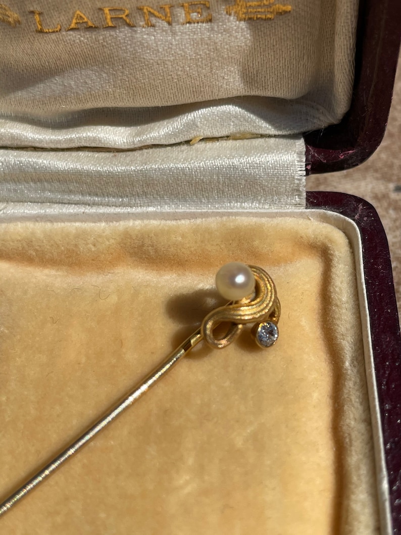May include: A gold-toned stick pin with a pearl and a small diamond-like stone. The pin is displayed inside a velvet-lined jewelry box with the word "LARNE" embossed on the inside of the lid.