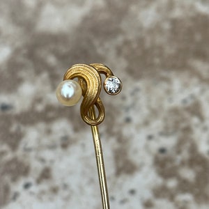 May include: A gold-toned stick pin featuring a swirling design with a small, round pearl on one end and a sparkling diamond on the other. The pin is slender and elegant, suitable for various occasions.