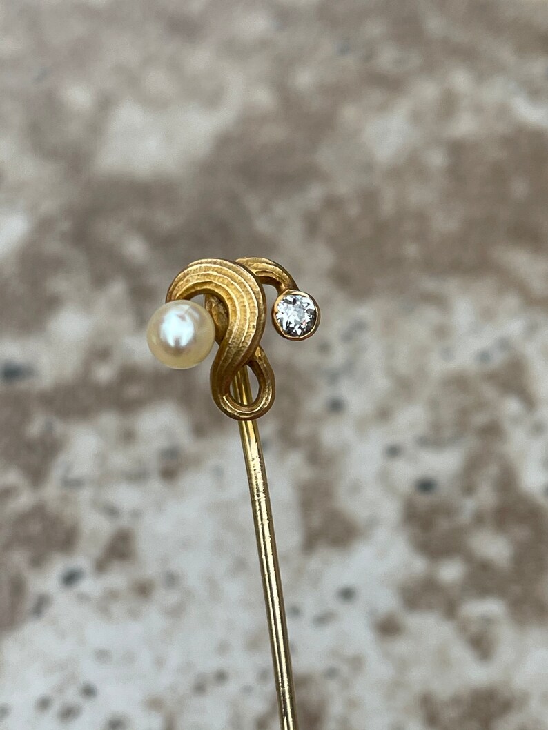 May include: A gold-toned stick pin featuring a swirling design with a small, round pearl on one end and a sparkling diamond on the other. The pin is slender and elegant, suitable for various occasions.