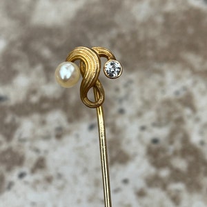 May include: A gold-toned hat pin with a swirling design. The pin features a small, round pearl on one end and a clear, round gemstone on the other. The pin is slender and has a pointed end.