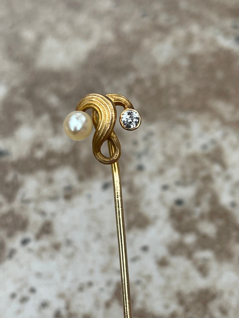 May include: A gold-toned hat pin with a swirling design. The pin features a small, round pearl on one end and a clear, round gemstone on the other. The pin is slender and has a pointed end.