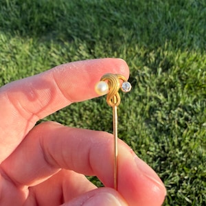 May include: A gold-colored hat pin with a pearl and a small, clear gemstone. The pin has an ornate, swirling design near the top. The background is blurred green grass.