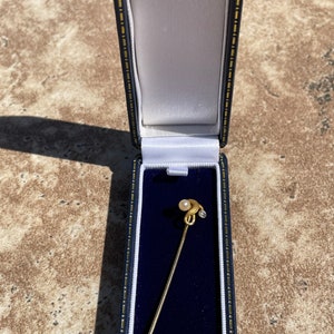 May include: A gold-colored stick pin with a pearl and a small diamond-like accent, presented in a blue and white lined jewelry box. The pin has a decorative, elongated head and a textured base.