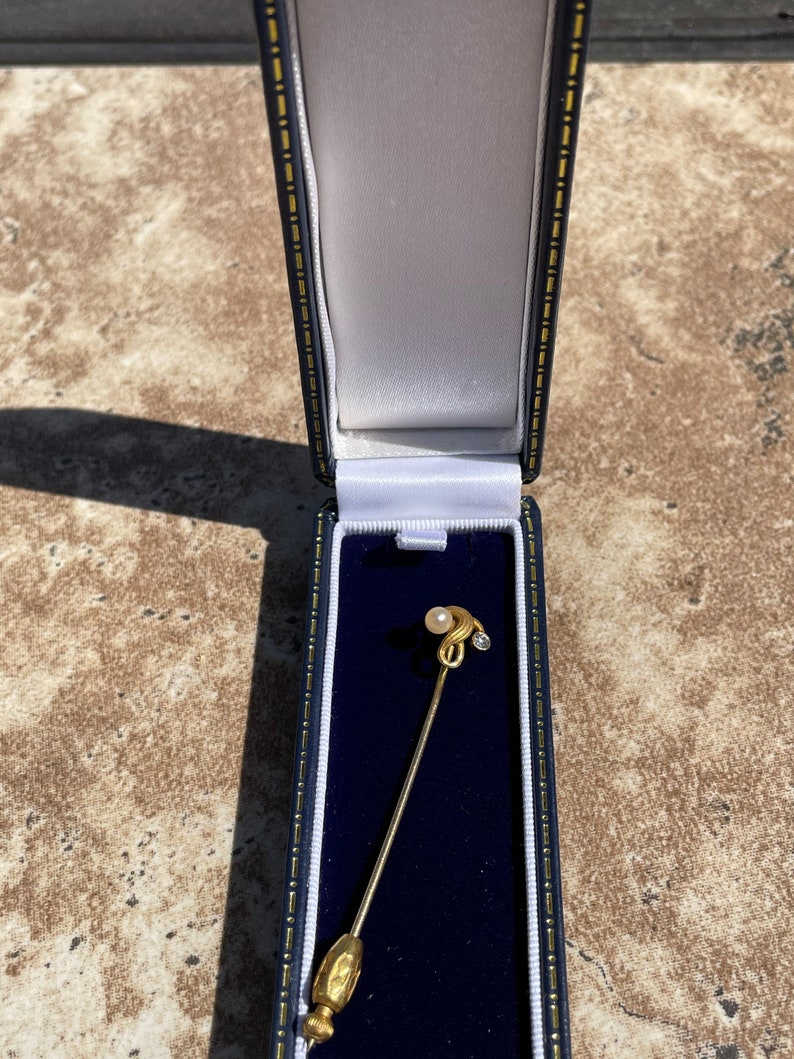 May include: A gold-colored stick pin with a pearl and a small diamond-like accent, presented in a blue and white lined jewelry box. The pin has a decorative, elongated head and a textured base.
