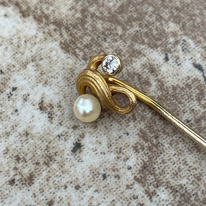 May include: A gold-toned stick pin with a pearl and a clear, round gemstone. The pin has a decorative, swirling design near the top, with the pearl at the bottom of the swirl. The gemstone is set above the swirl.