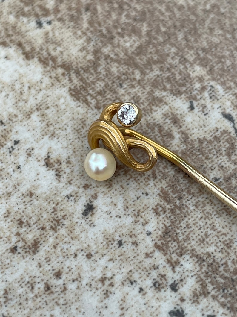 May include: A gold-toned stick pin with a pearl and a clear, round gemstone. The pin has a decorative, swirling design near the top, with the pearl at the bottom of the swirl. The gemstone is set above the swirl.