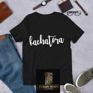 Könnte beinhalten: Schwarzes T-Shirt mit dem Wort "bachatera" in weißer Kursivschrift. Das Shirt hat ein kleines Logo unten, das "Turbo Body - USA Corp." lautet.