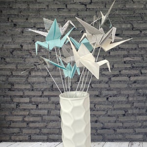 May include: A white ceramic vase with a geometric pattern holds a bouquet of origami paper cranes. The cranes are in shades of white, light blue, and gray, some with patterned designs. The vase sits on a white wooden surface against a gray brick wall.