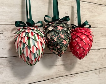 May include: Three handmade paper Christmas ornaments in the shape of pine cones. The ornaments are decorated with different patterns: red and green stripes, floral print, and red with white stars.