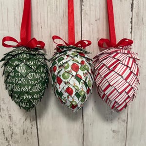 May include: Three decorative Christmas ornaments shaped like pinecones, each suspended by a red ribbon. One is green with white text, another has a pattern of red and green ornaments, and the third has red and white stripes.