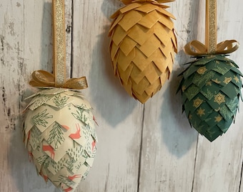 May include: Three paper pinecone ornaments with gold ribbon. The pinecones are decorated with different patterns: one is solid gold, one is green with snowflakes, and one is white with a green and pink floral pattern.