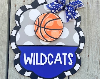 May include: A gray and white polka dot door hanger with a blue and white bow. The door hanger has a basketball and the word "WILDCATS" in white letters on a blue background.