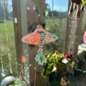 May include: A clear, iridescent, plastic UFO charm with a small alien figure hanging from it. The charm is attached to a gold-colored wire.