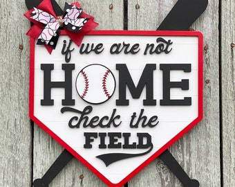 May include: A white wooden home plate sign with red trim and black lettering. The sign says "If we are not home check the field". A baseball is centered on the sign. Two black wooden baseball bats are crossed behind the sign. A red and white polka dot bow with a black and white baseball print is attached to the top of the sign.