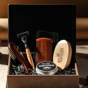 Könnte beinhalten: Ein Herren-Pflegeset in einer schwarzen Box. Das Set enthält eine Holzbürste, einen Rasierer, einen Kamm, einen Bart- und Schnurrbartbalsam und eine ovale Holzbürste mit dem Text "NICOLAS".