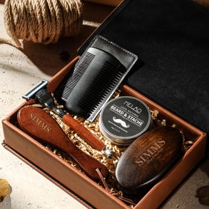 Könnte beinhalten: Ein braunes Lederbox-Set mit Pflegeutensilien. Das Set enthält einen Kamm, einen Rasierer, eine Bartbürste und eine Dose Bart- und Schnurrbartbalsam. Die Box ist geöffnet und zeigt den Inhalt vor einem neutralen Hintergrund.