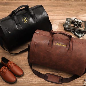 Könnte beinhalten: Eine schwarze Reisetasche mit goldenem K-Monogramm und eine braune Reisetasche mit dem gold gestickten Namen Nathaniel. Ebenfalls enthalten sind ein Paar braune Schnürschuhe, eine Kamera, ein Buch mit dem Titel AB CONCEPT und eine silberne Armbanduhr.