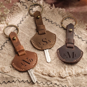 Könnte beinhalten: Drei Leder-Schlüsselanhänger mit Messingringen und personalisierten Initialen. Die Schlüsselanhänger sind in Brauntönen gehalten und mit den Initialen "AND", "VSN" und "KML" graviert. Die Schlüssel sind sichtbar, der Hintergrund ist ein Stoff.